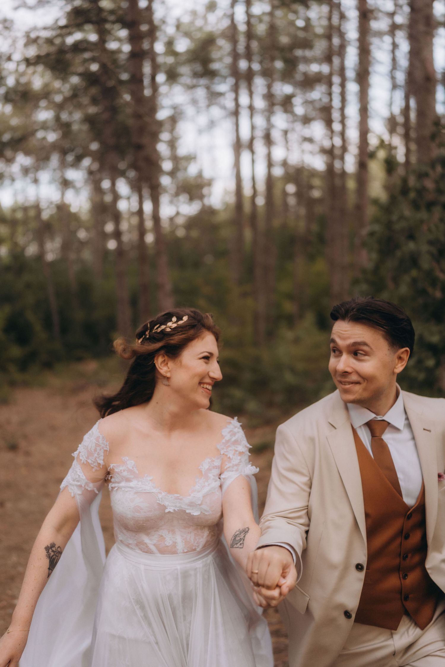 Bride and groom hold hands smiling at each other while walking through a pine forest in wedding attire.