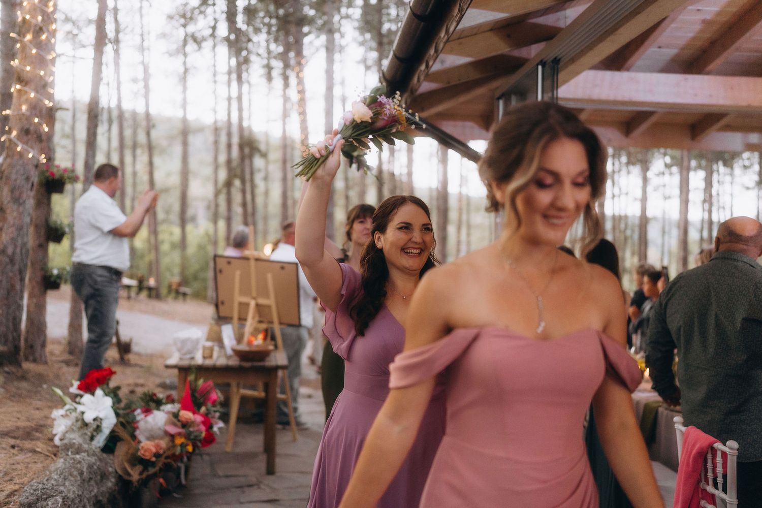 Bridesmaids in mauve dresses celebrate at an outdoor woodland wedding reception with string lights and pine trees.