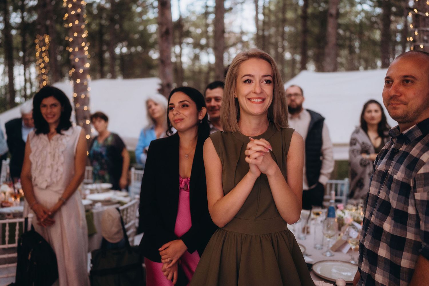 Smiling blonde wedding guest in olive dress clasps hands joyfully at an outdoor forest reception.
