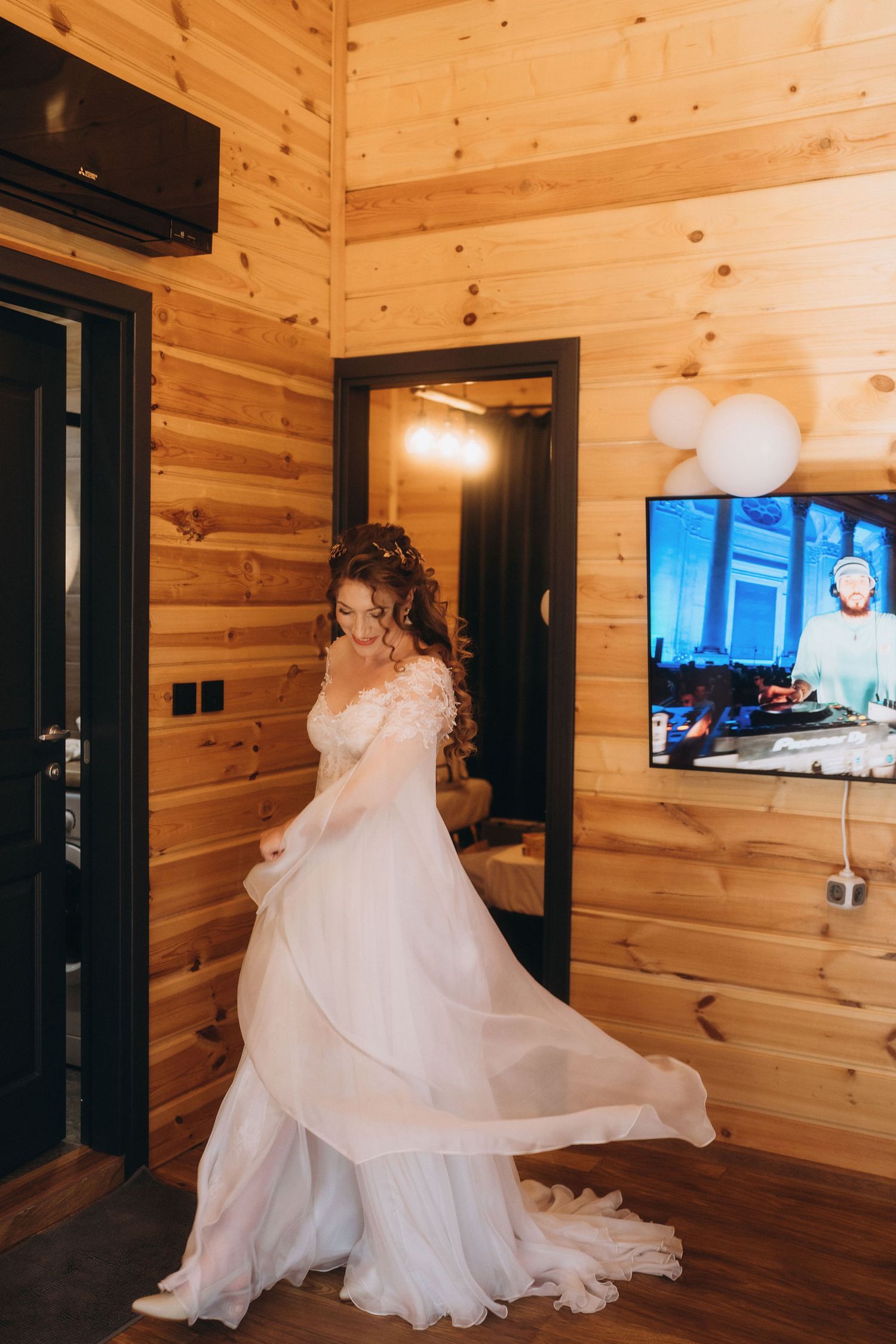 Bride in elegant white wedding gown twirling in a wooden cabin room with TV and mirror.