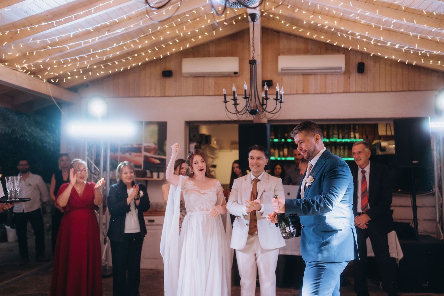 Bride and groom celebrate with champagne at elegant wedding reception with string lights and chandelier.