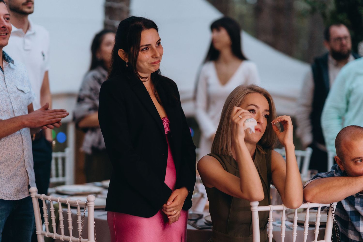Emotional wedding guests watch ceremony, woman wiping tears at outdoor reception event.