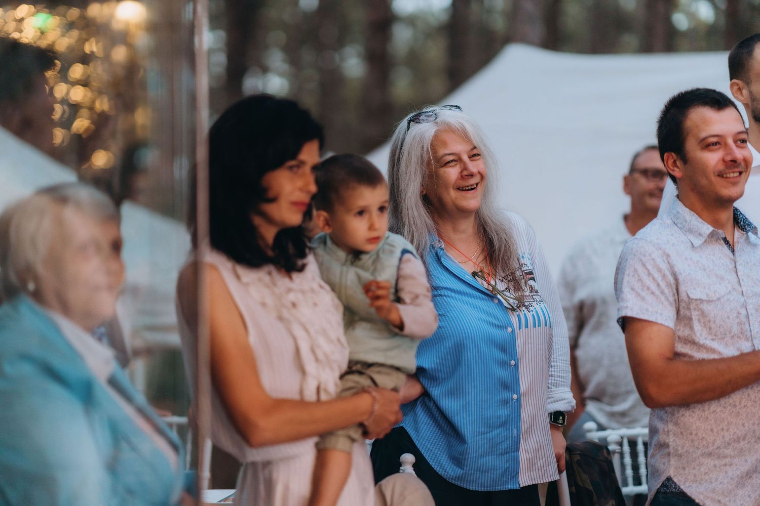 Smiling wedding guests including a woman holding a baby and an older woman with gray hair enjoy an outdoor ceremony.