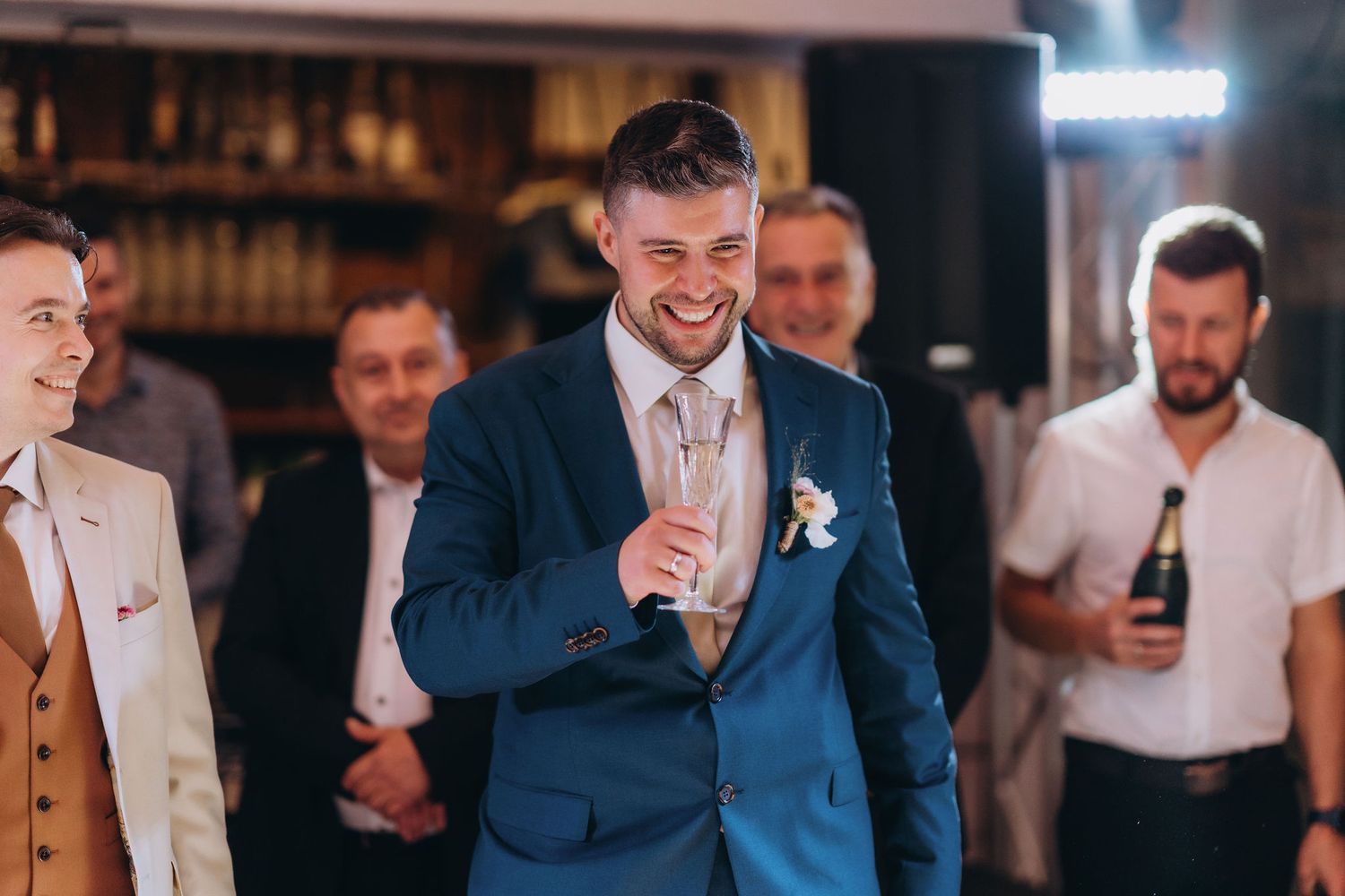 Smiling groom in blue suit holding champagne glass with groomsmen laughing in background at wedding reception.