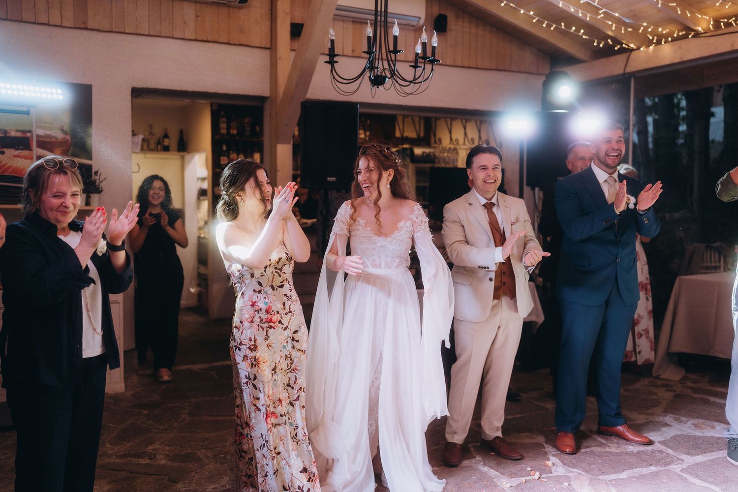 Bride in flowing white lace gown enters wedding reception hall with guests clapping and celebrating.