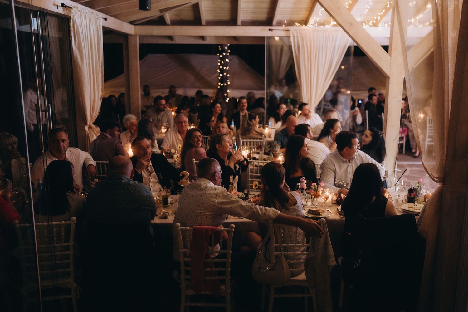 Large wedding reception dinner with guests seated at candlelit tables in decorated venue.