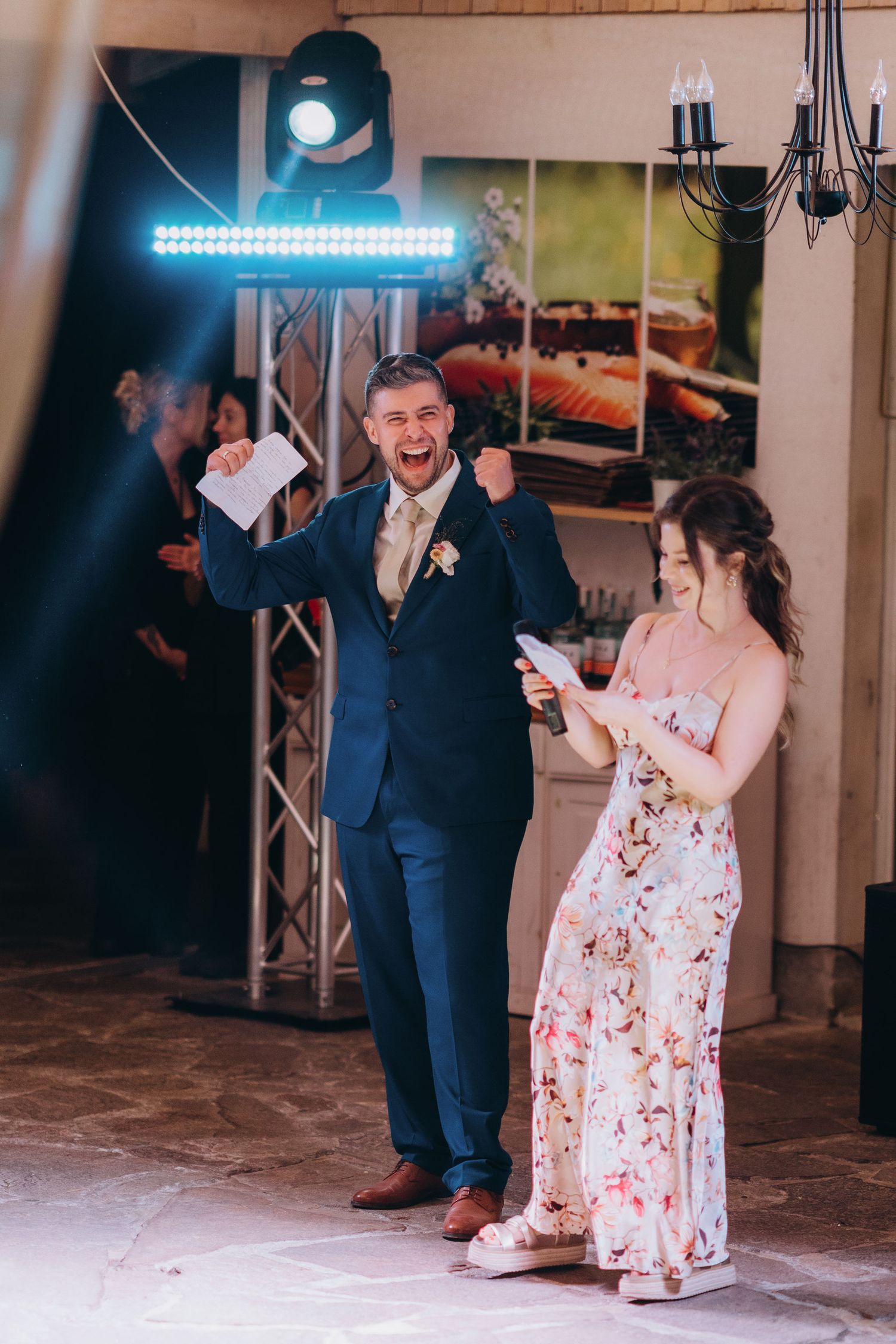 A groom in a navy suit celebrates with a woman in a floral dress during a wedding reception speech indoors.