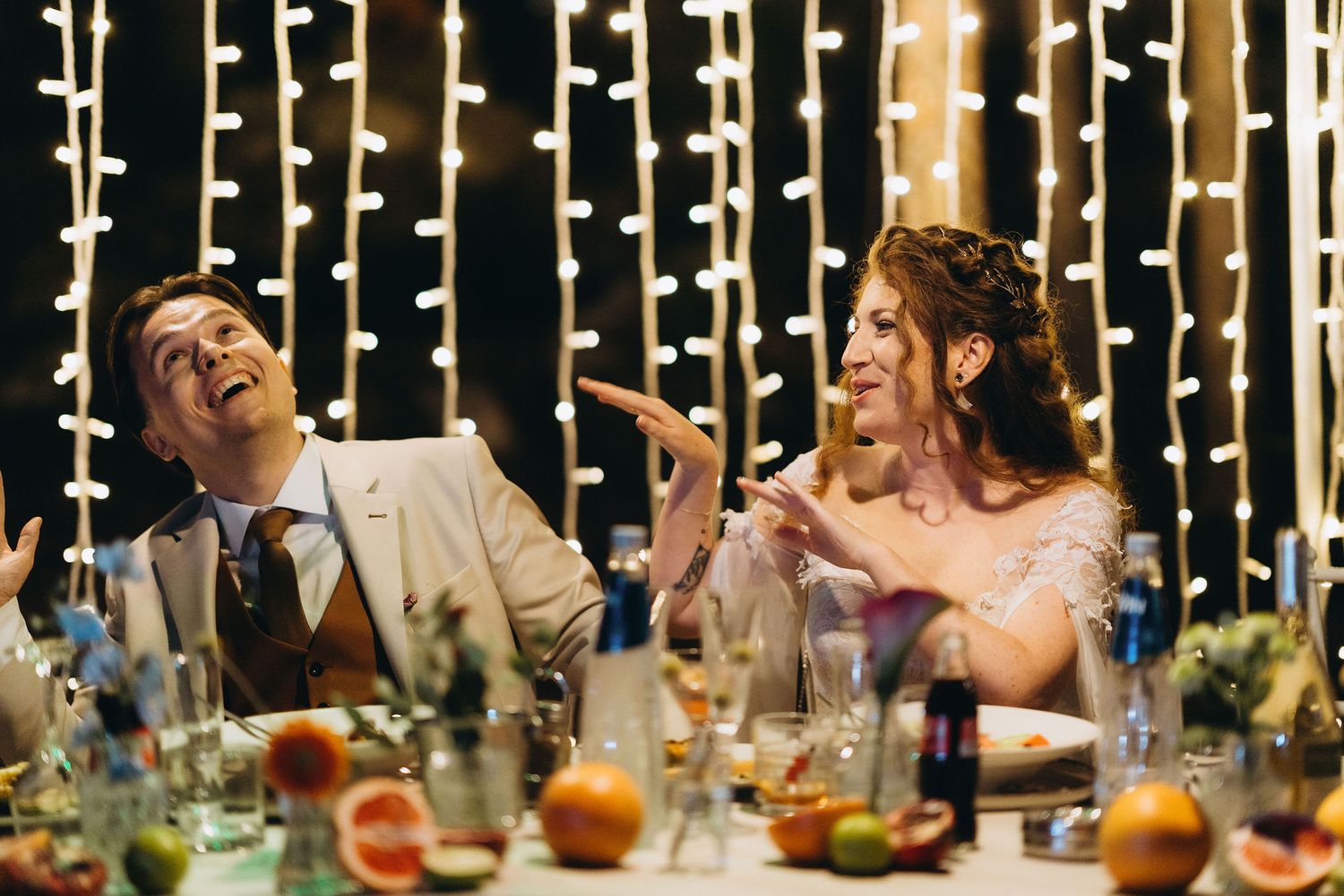 Bride and groom laugh joyfully at wedding reception table adorned with fairy lights and fresh fruit.