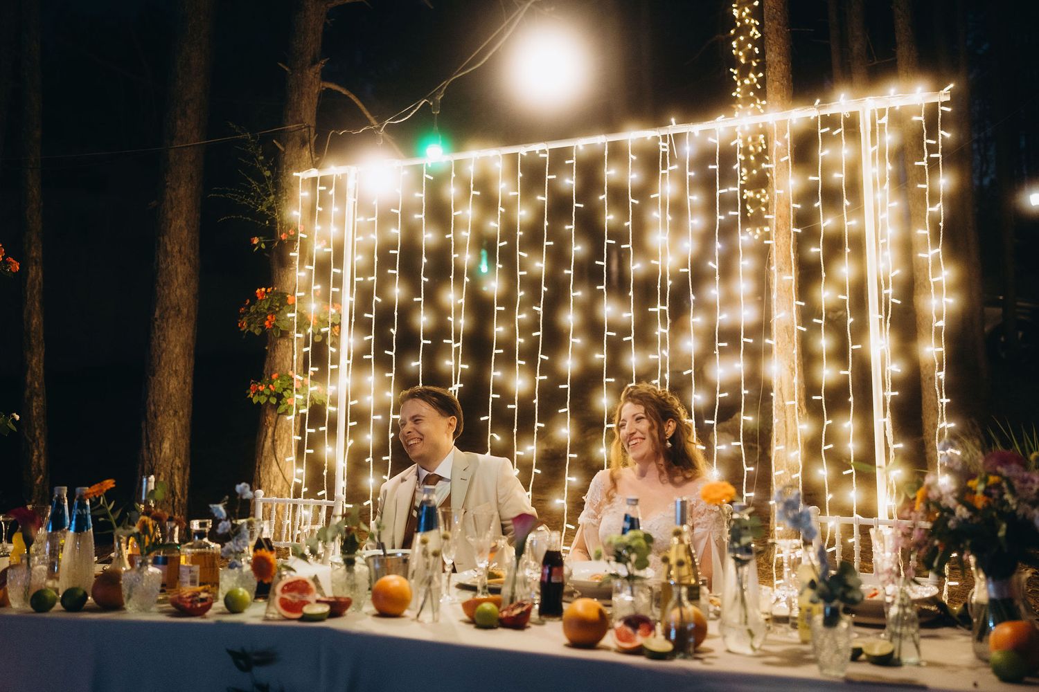 Bride and groom seated at elegant outdoor wedding table adorned with fairy lights and floral arrangements at night.