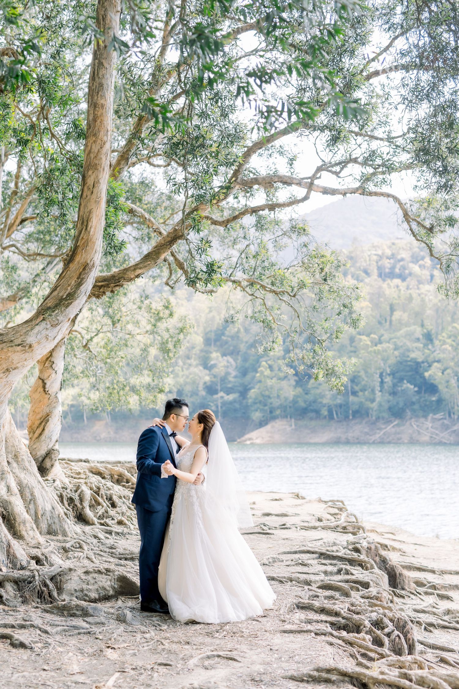 Bride and groom embracing under a large tree by a serene lake, surrounded by lush greenery on their wedding day.