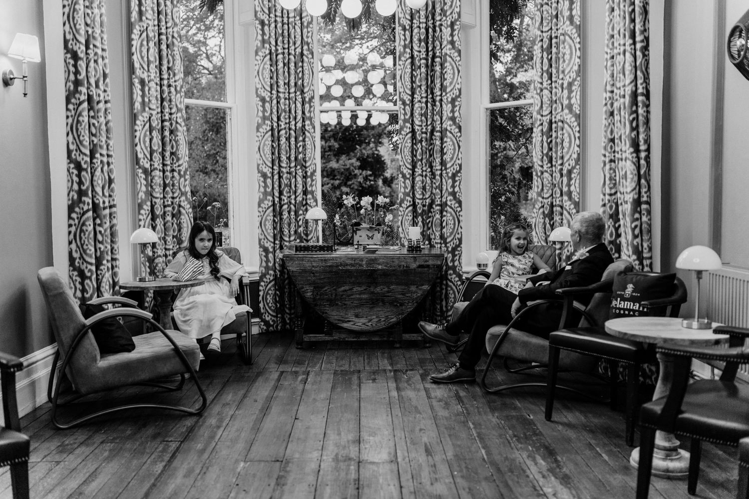 Black and white photo of people relaxing in a vintage living room with patterned curtains and hardwood floors.