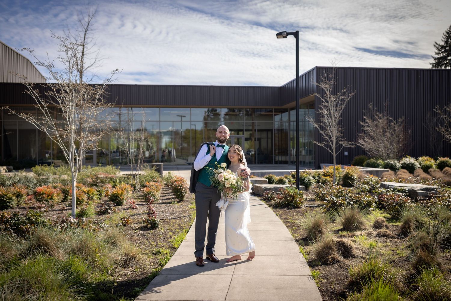 Bride and groom pose on a garden pathway in front of a modern building.