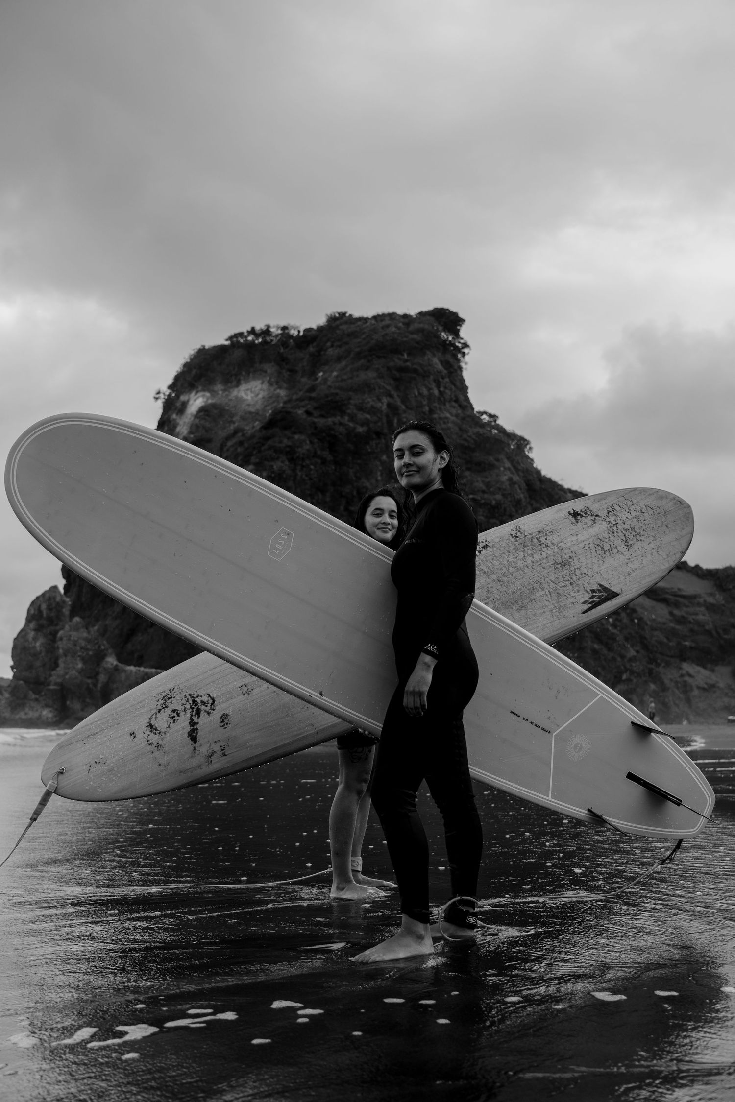 photo of two diverse women surfers posing at the Lion Rock at Piha, Auckland Aotearoa New Zealand