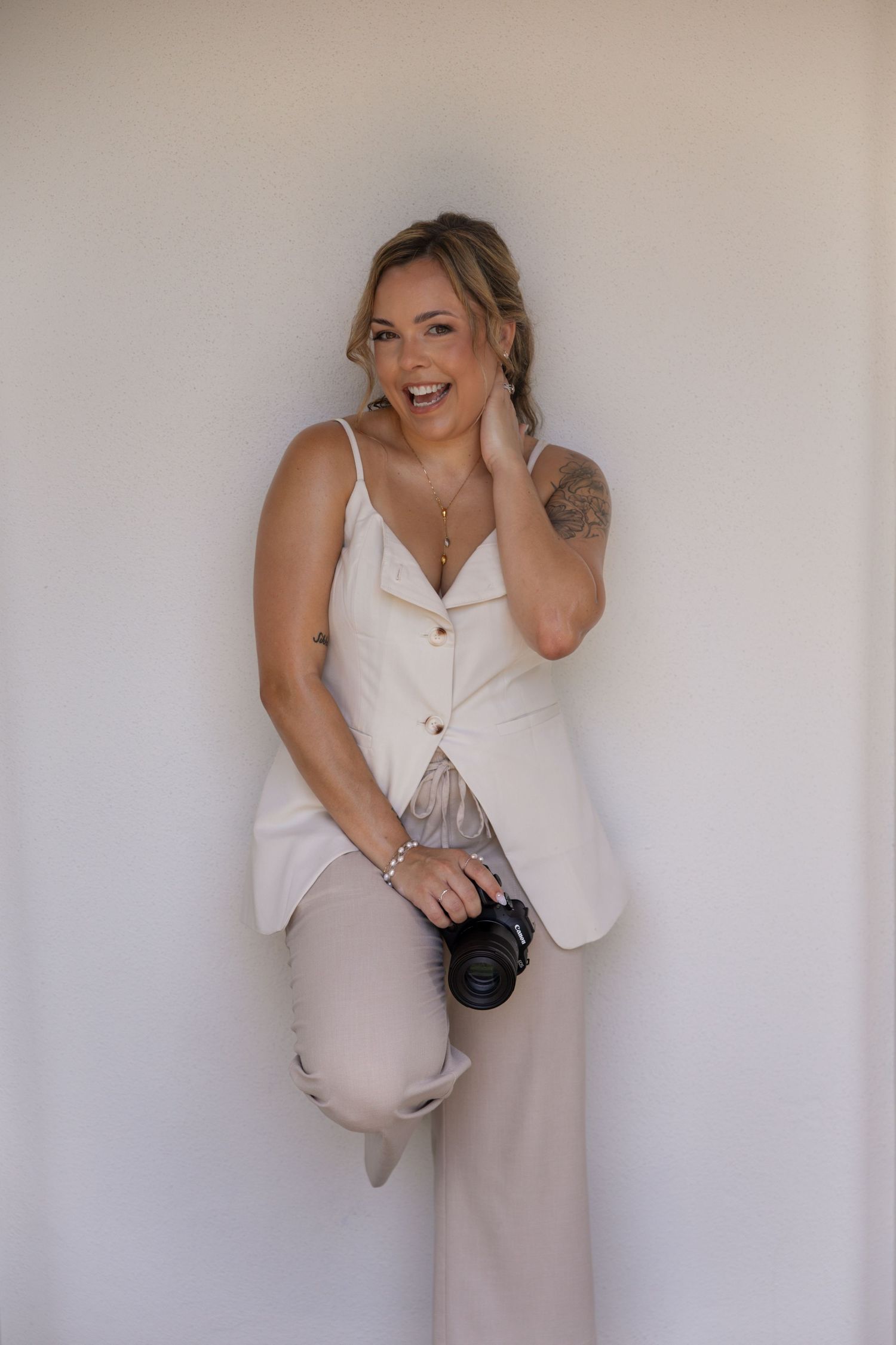 Smiling woman in a cream vest and wide-leg trousers leans against a white wall, holding a camera with one knee raised.