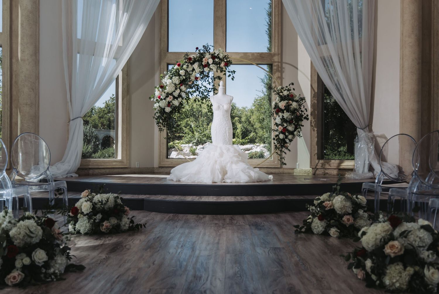 lace mermaid wedding dress on a mannequin in front of a circular floral arch with white and red roses and modern ghost chairs.
