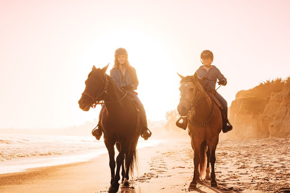 Two riders on horseback walking along a sunny beach at golden hour with ocean waves and cliffs in background.