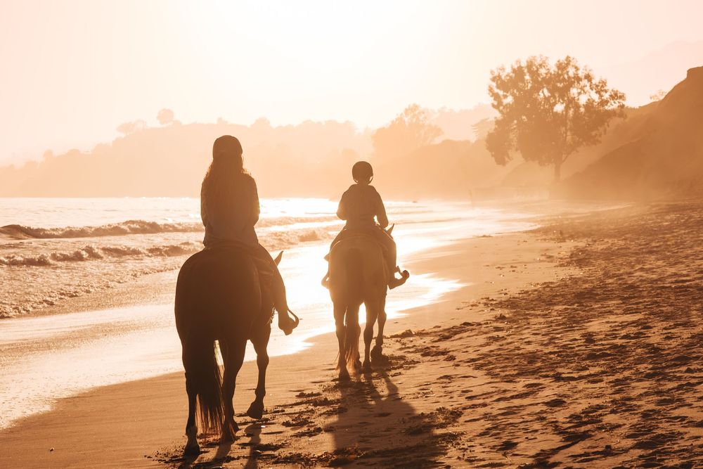 Two riders on horseback walking along a sandy beach at golden sunset with warm glowing light and trees in background.