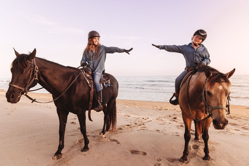 Two riders on horseback pose with outstretched arms on a sandy beach at sunset, wearing helmets and casual attire.