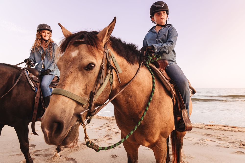 Two women riding brown horses on a sandy beach at sunset, wearing helmets and casual clothing.