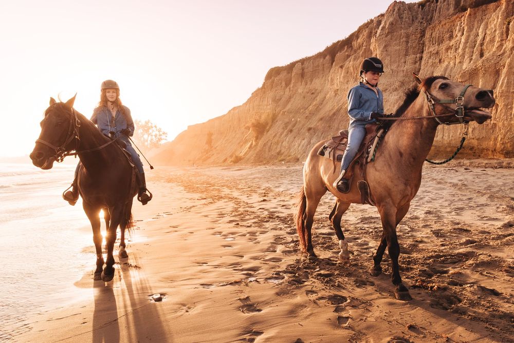 Two riders on horseback along a sandy beach at golden sunset with dramatic cliffs in the background.