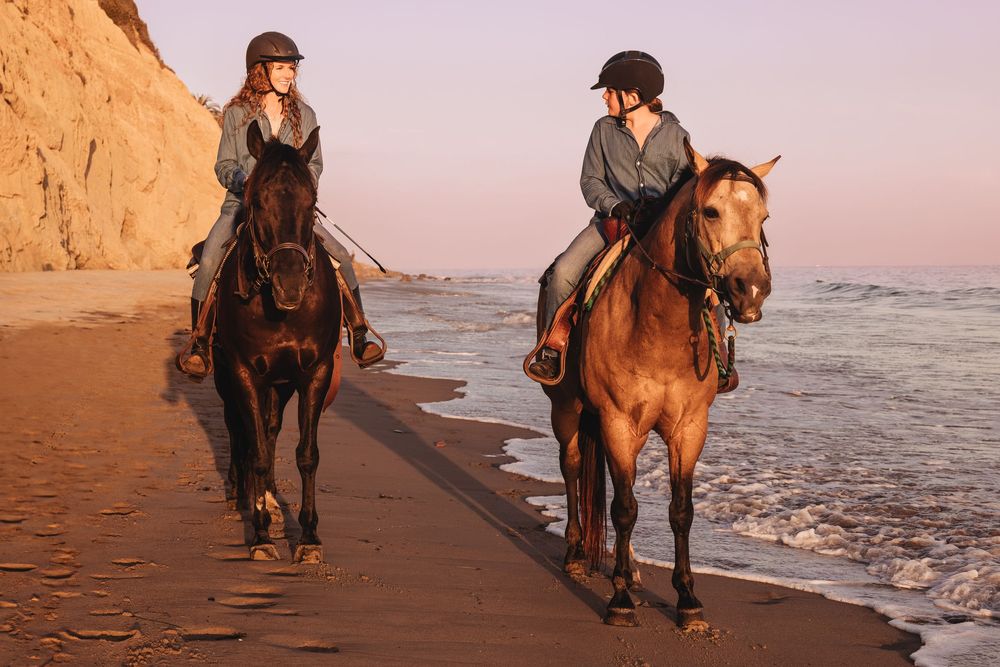 Two riders on horseback walk along a sandy beach at sunset near ocean waves.