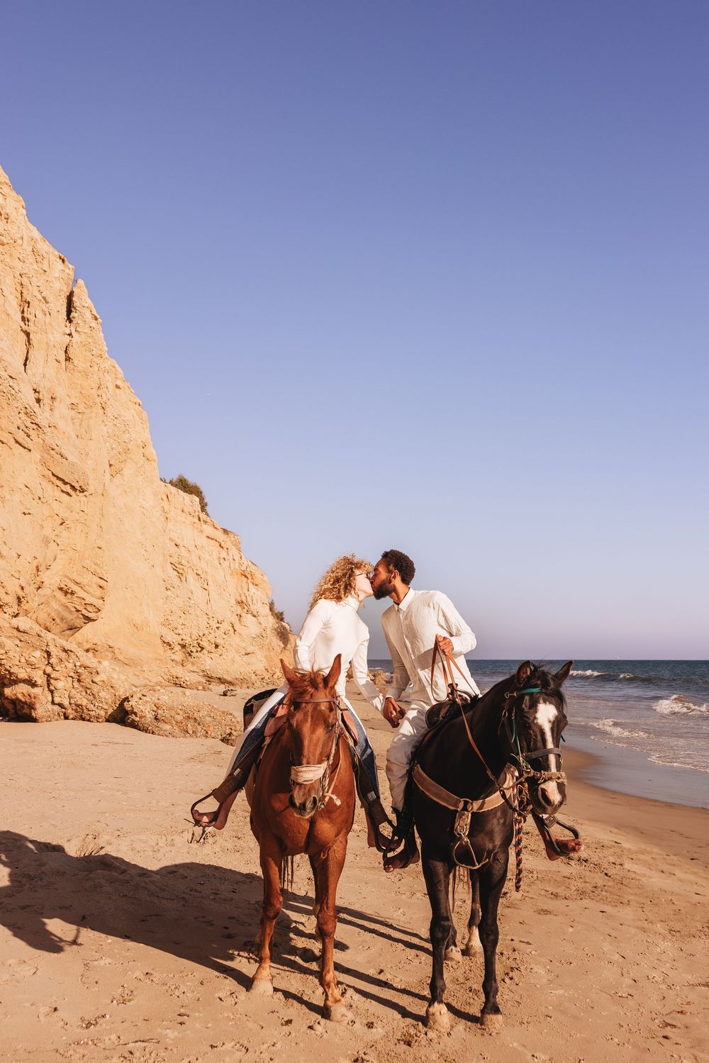 Romantic couple kissing on a sunny beach while holding hands beside two horses near golden cliffs.