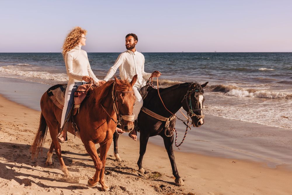 Couple riding horses together on a sandy beach beside ocean waves on a sunny day.