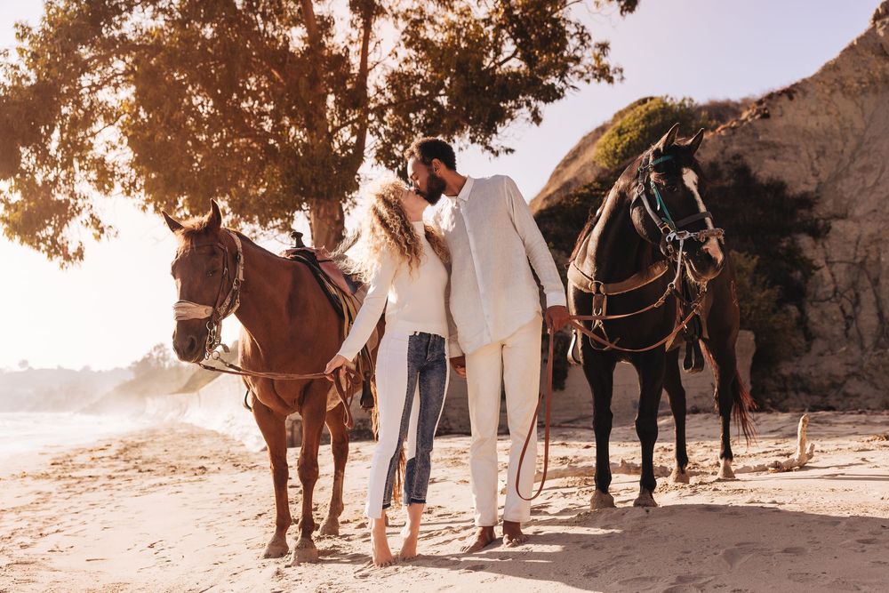 Couple walking with two horses on a sunny beach, dressed in elegant white and casual attire.