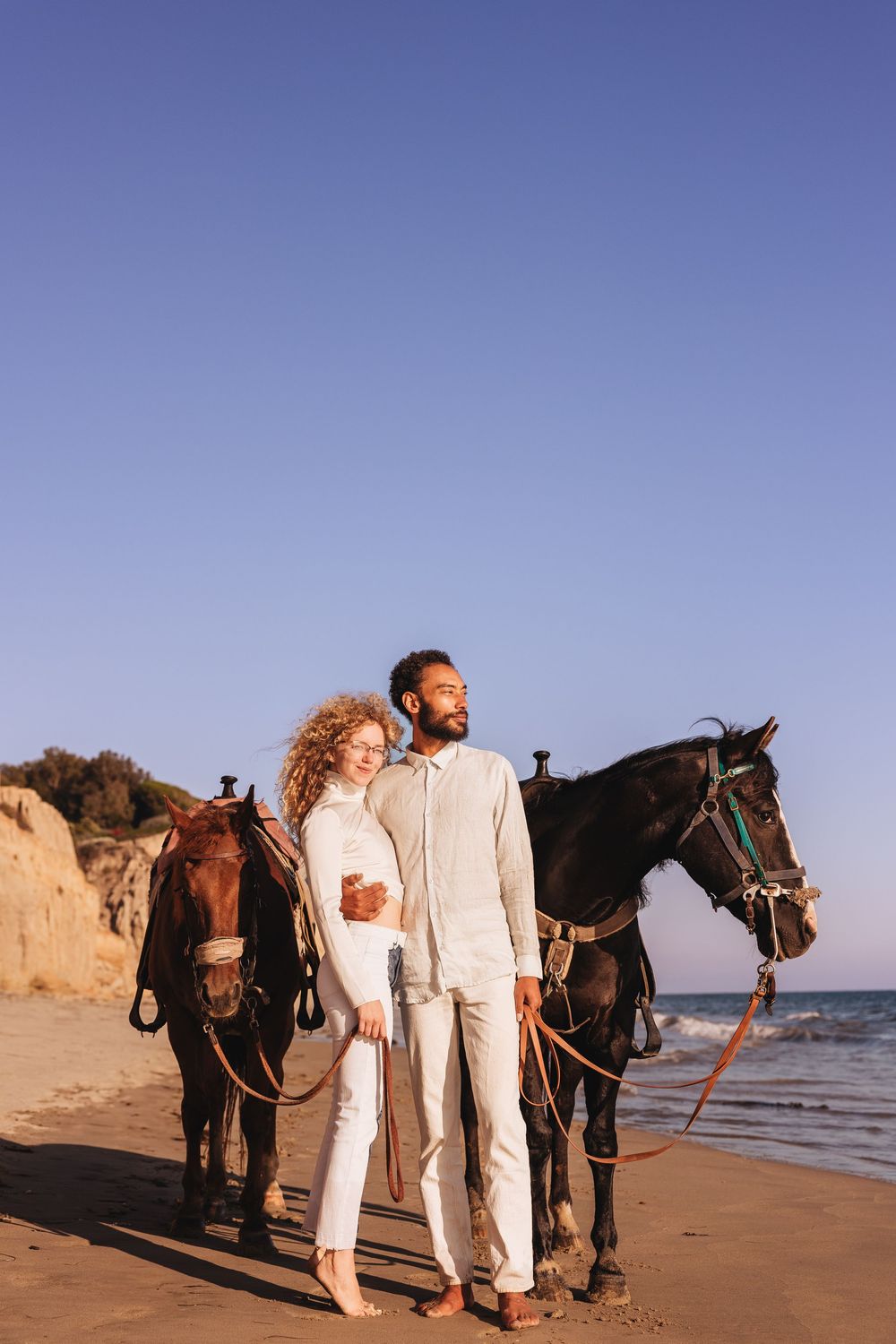 Couple standing together with two horses on a sandy beach at sunset near rocky cliffs.
