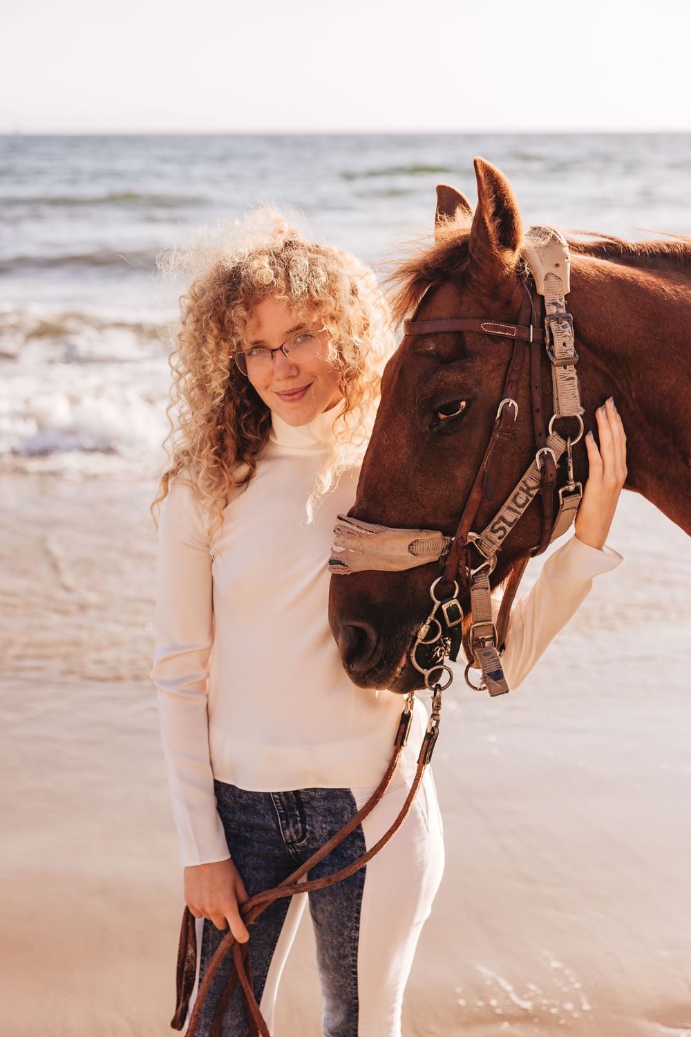 A curly-haired woman in a white sweater stands with a brown horse on a sunny beach near the ocean.