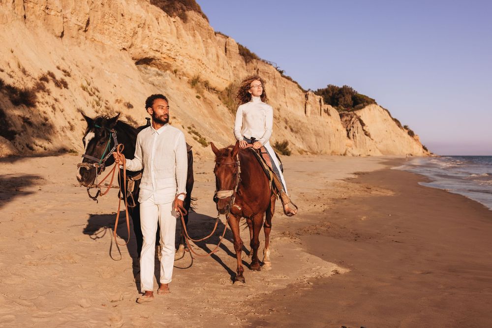 Couple walking with horses on a sunny beach near sandy cliffs, man in white suit leading brown horse.