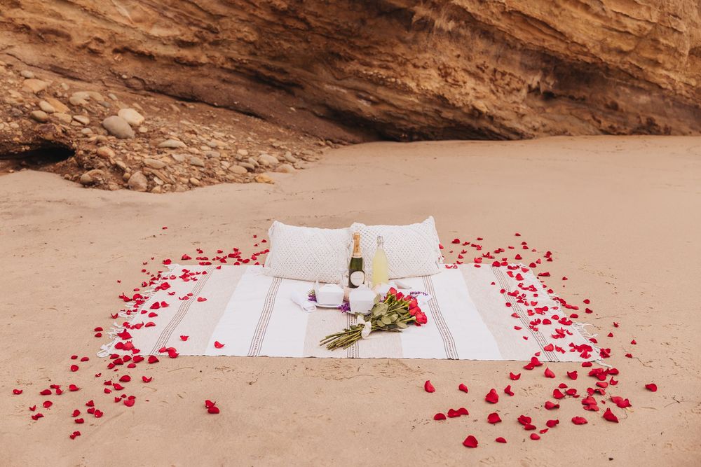 Romantic beach picnic setup with rose petals, champagne, and flowers on white blanket against sandy cliffs.