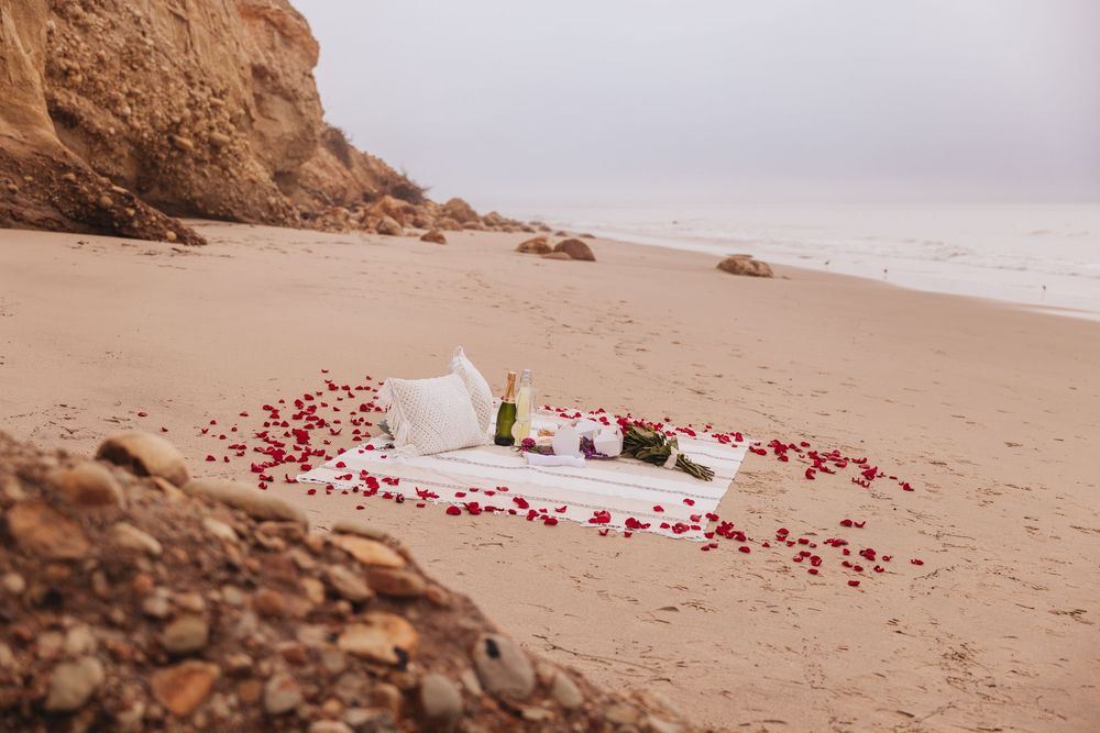 Romantic beach picnic setup with rose petals, champagne, and white blanket on sandy shore near rocky cliffs.