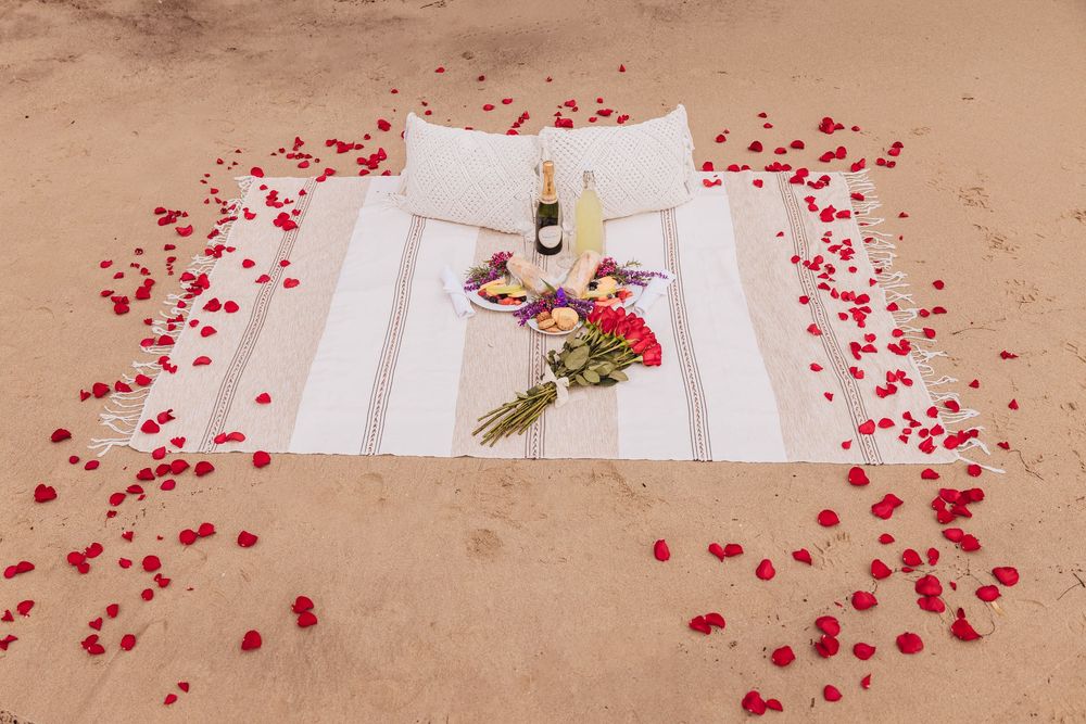 Romantic beach picnic setup with a striped blanket, bouquet, champagne bottle, and red rose petals scattered on sand.
