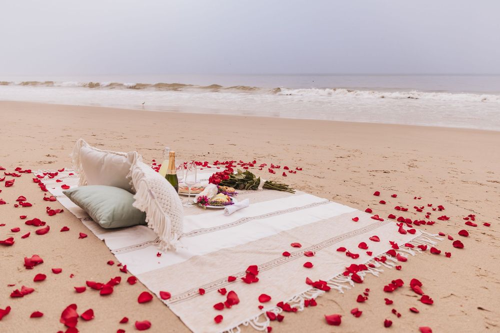 Romantic beach picnic setup with red rose petals scattered on sand, blanket, cushions, and champagne by the ocean.