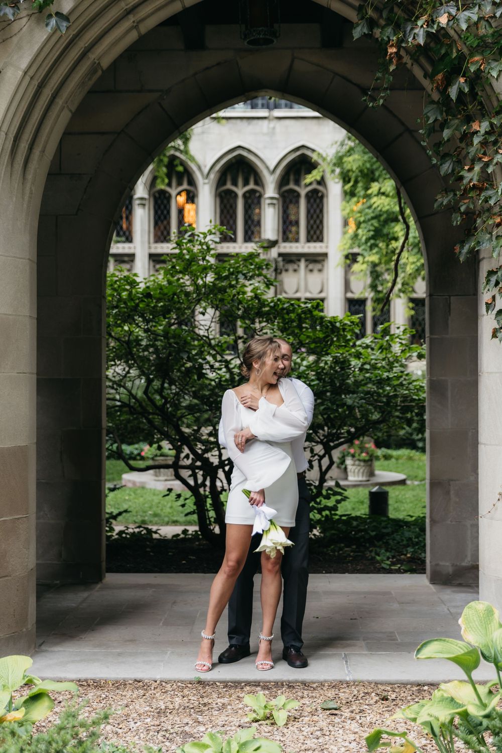 Couple embracing under stone arch in wedding attire, bride in white mini dress holding bouquet, Chicago fourth presbyterian church backdrop