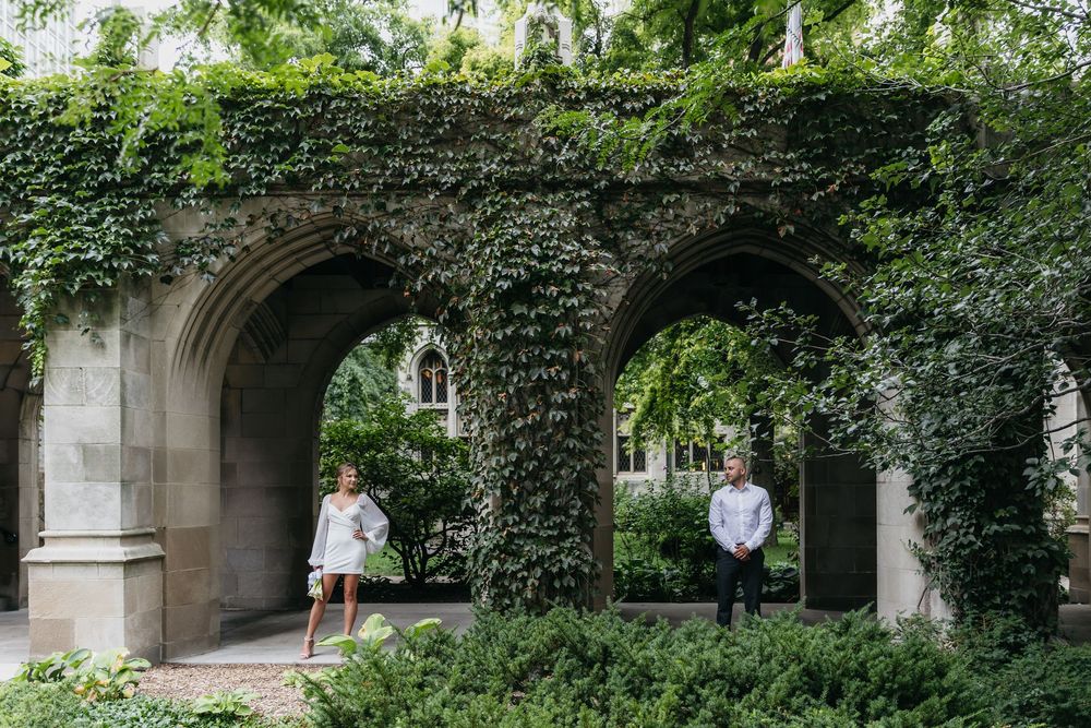 Couple standing under stone arches in wedding attire, bride in white mini dress holding bouquet, Chicago fourth presbyterian church backdrop