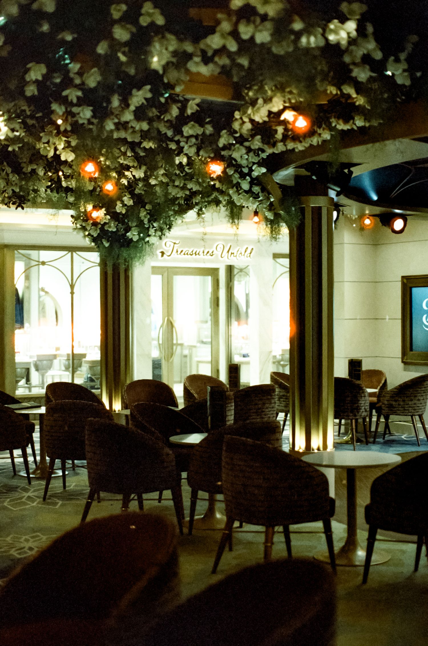 Elegant restaurant interior with floral ceiling decor, warm string lights, and dark wicker chairs around marble tables.