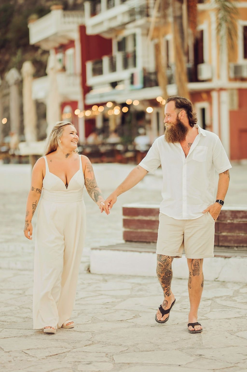 Tattooed couple holding hands and walking together in white outfits at a scenic coastal town plaza.