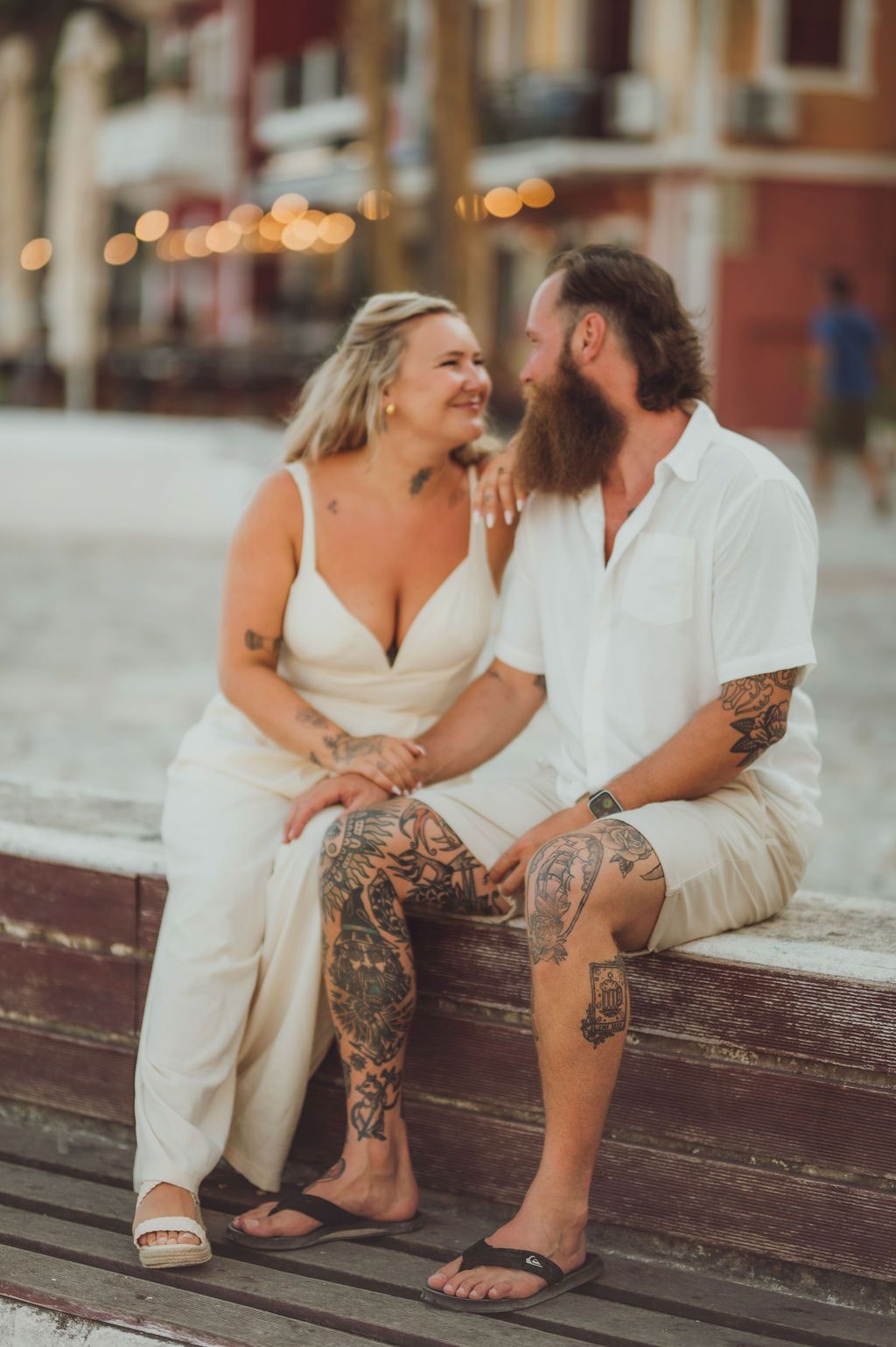 Tattooed couple in white outfits sitting on a wooden bench, smiling at each other in a warm outdoor setting.