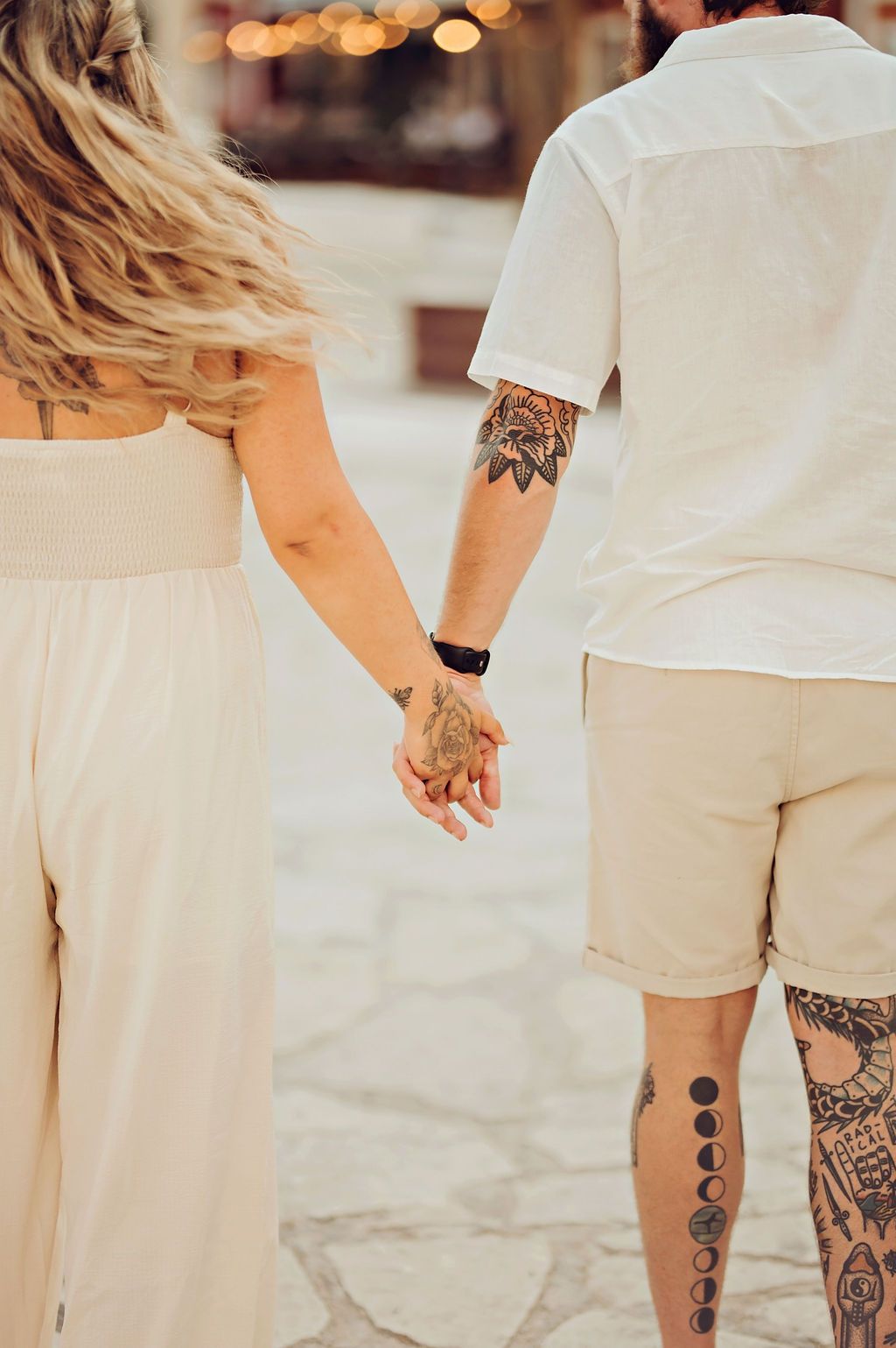 Couple holding hands while walking, both with tattoos, wearing neutral tones, shot from behind on cobblestone.