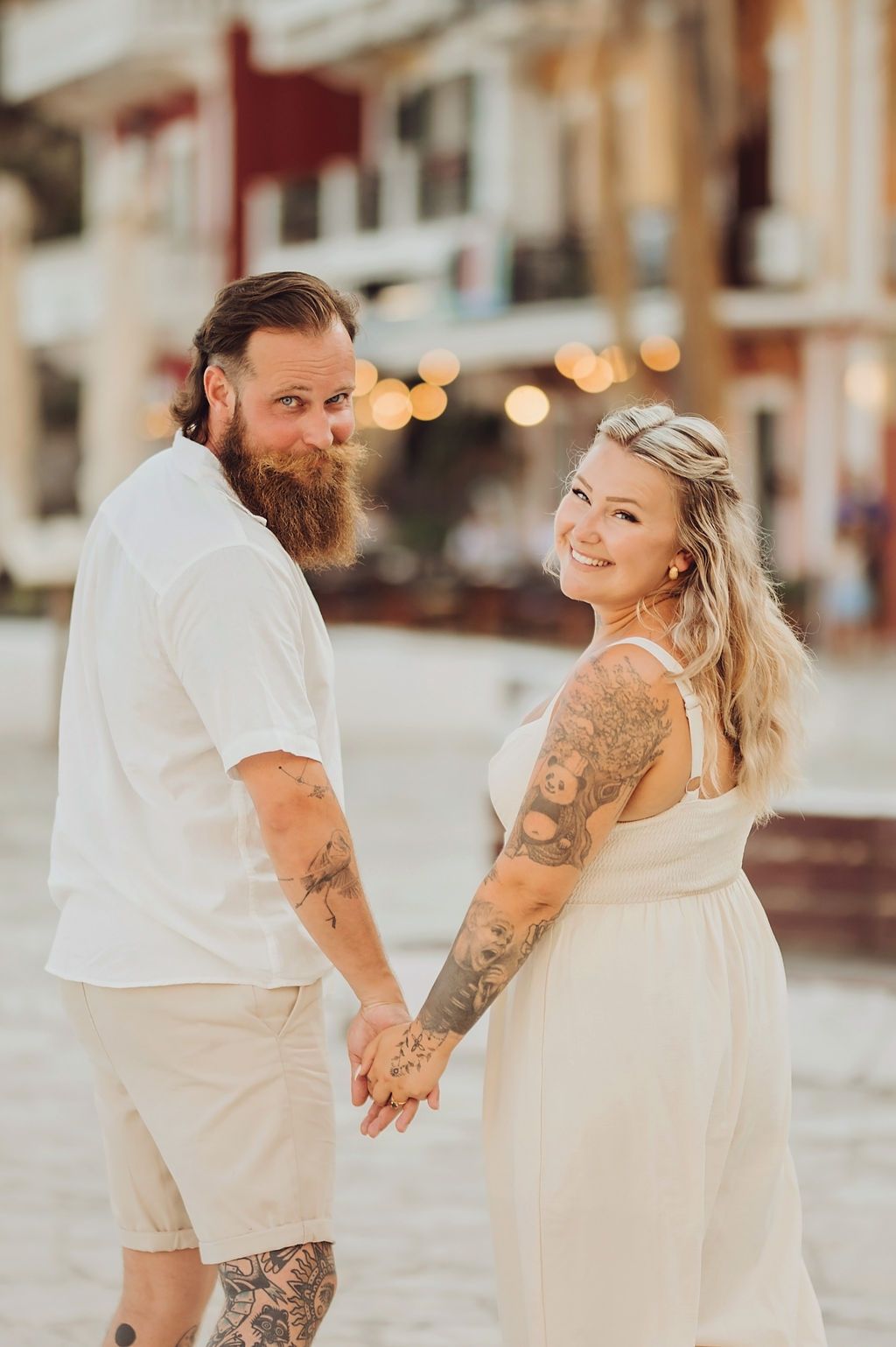 Tattooed couple in white outfits holding hands and smiling outdoors with bokeh lights in background.