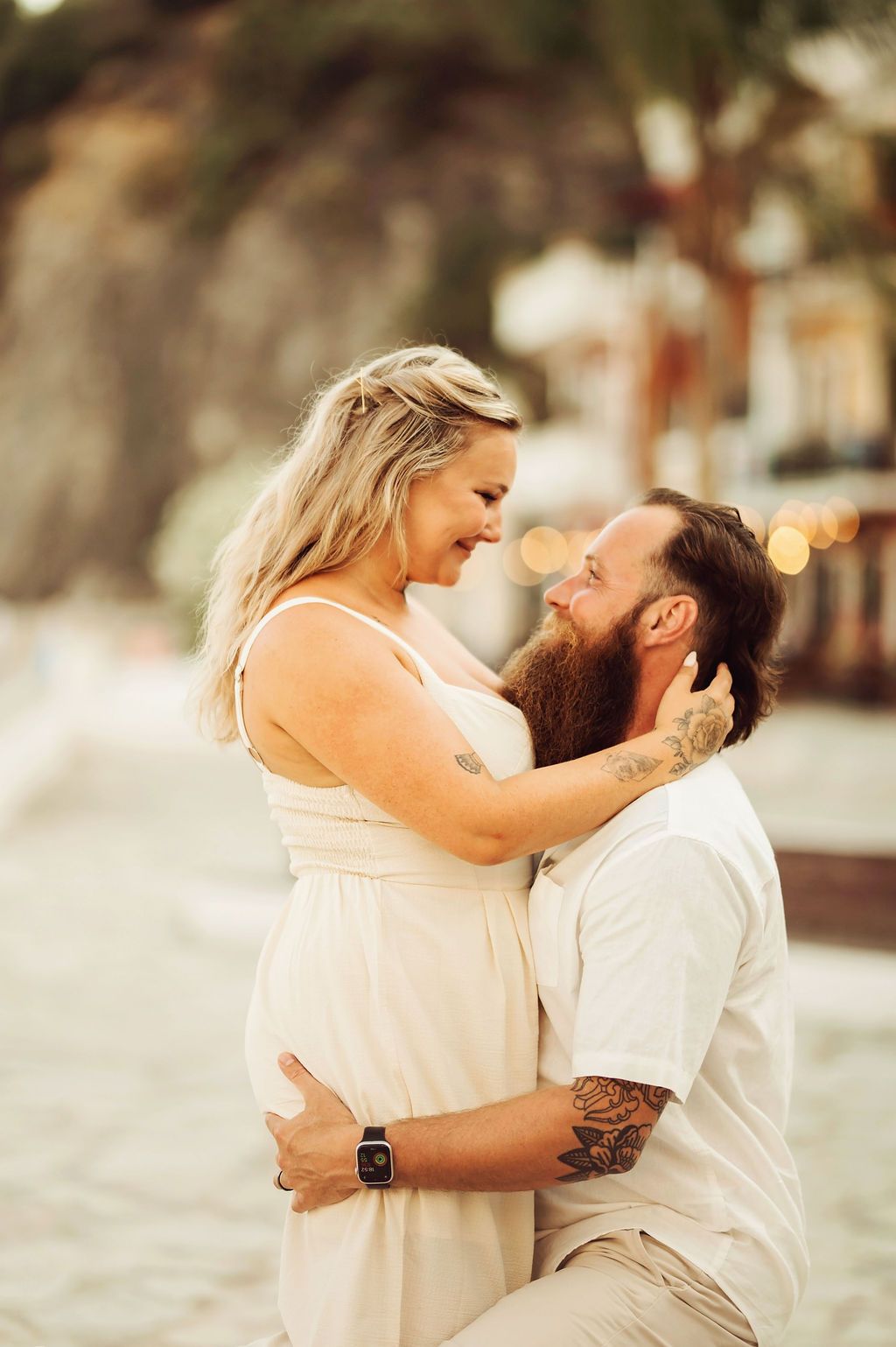 A bearded man with tattoos kneels before a smiling blonde woman in a white dress during a romantic beach maternity photo session.