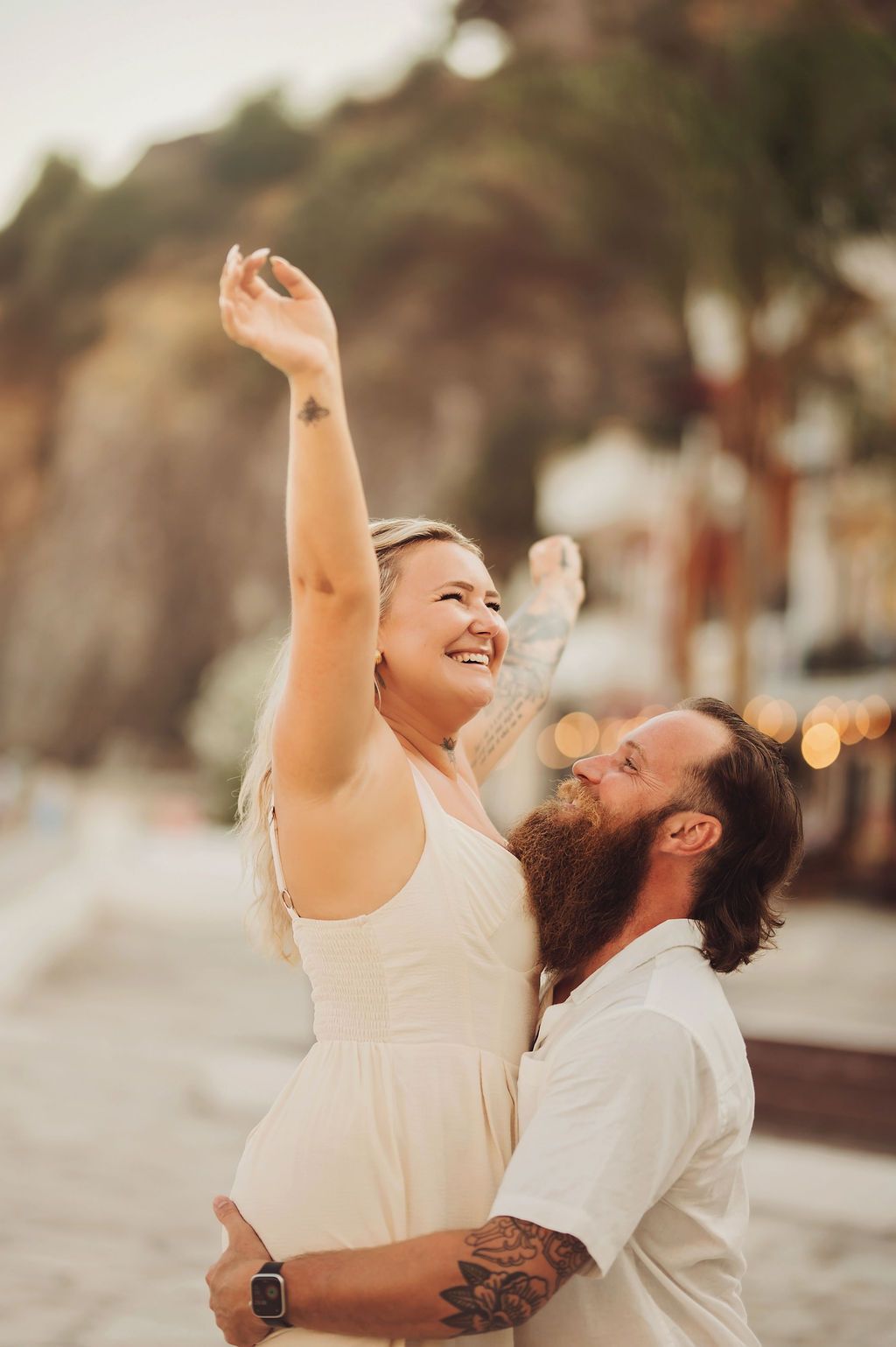 Joyful couple embracing on a beach, woman in white dress raising her arm, man with beard and tattoos, golden hour.