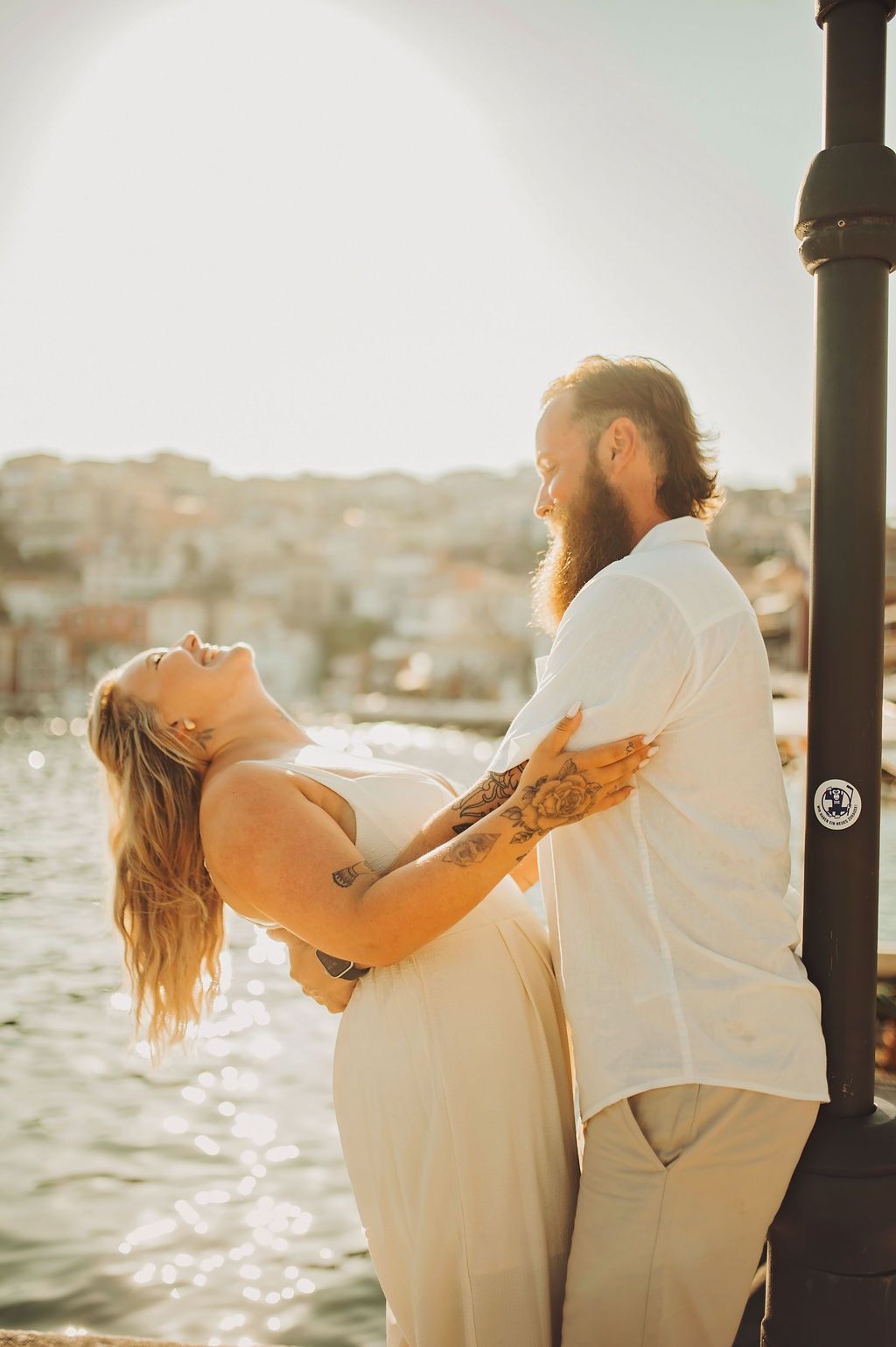 Romantic couple dancing by waterfront at sunset, woman leaning back laughing, bearded man with tattoos in white shirt.