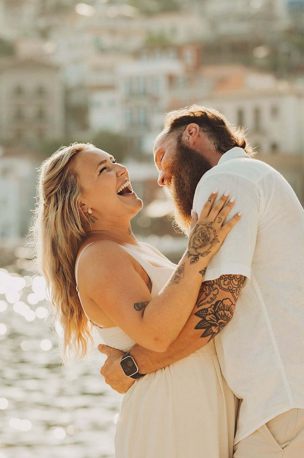 Laughing couple embracing outdoors with tattoos, white outfits, and a blurred coastal town background in warm sunlight.