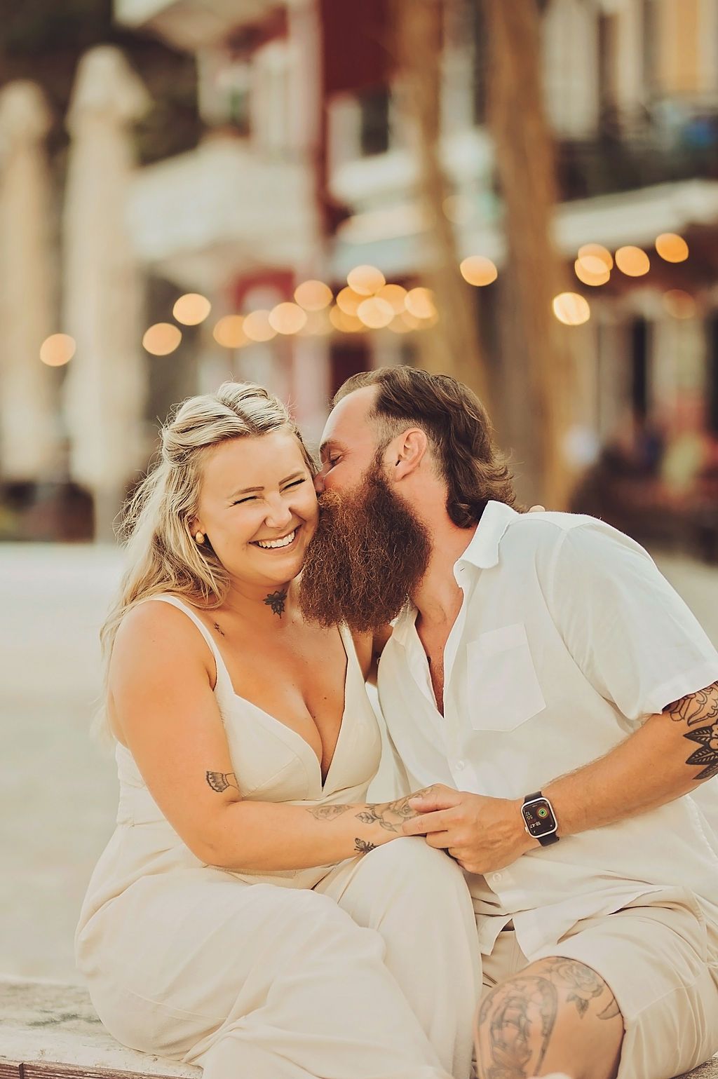 A smiling couple in white outfits with tattoos share a tender moment outdoors, with bokeh lights in the background.