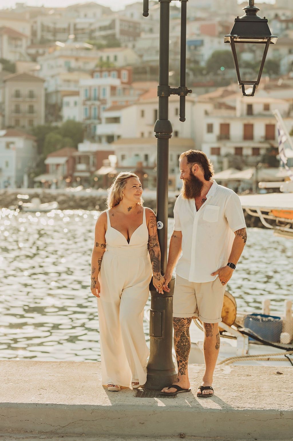 Tattooed couple holding hands by a lamppost on a Mediterranean waterfront promenade, dressed in white outfits.