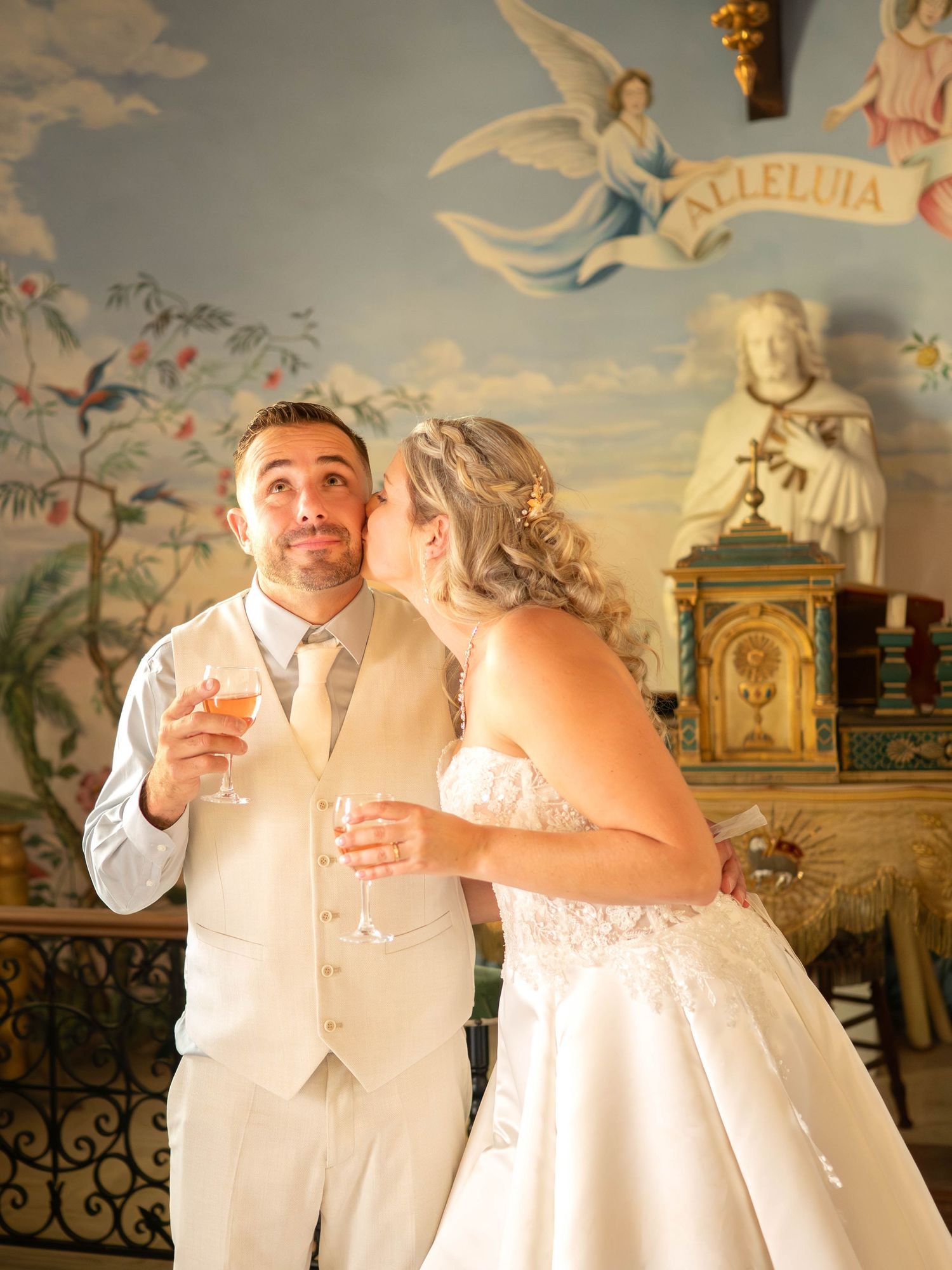 La mariée embrasse le marié sur la joue, un verre de champagne à la main, devant une fresque colorée d'une chapelle d'un Domaine