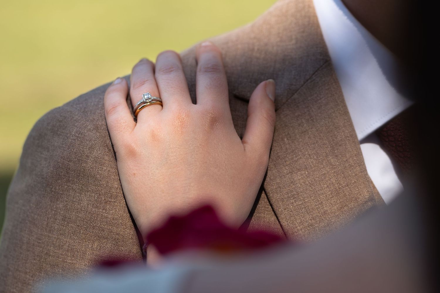 Close-up of bride's hand with gold wedding ring resting on groom's tan suit jacket.